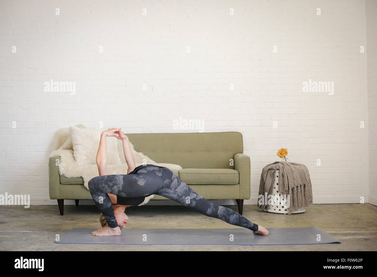 Una donna bionda in un nero leotard e gambali, in piedi su un materassino yoga in una stanza, fare yoga, la sua gambe divaricate e braccia alzate. Foto Stock