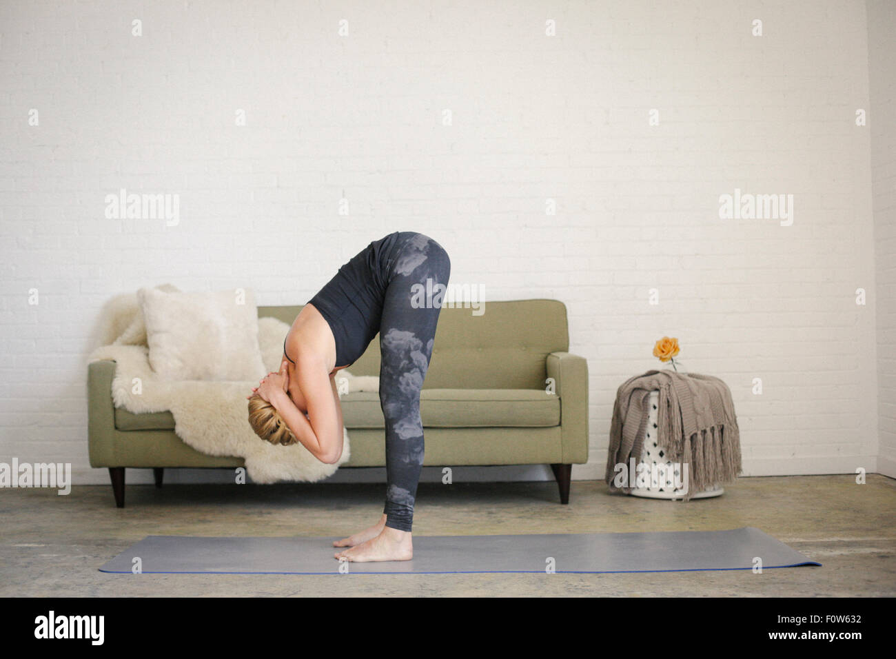 Una donna bionda in un nero leotard e gambali, in piedi su un materassino yoga in una stanza la flessione in avanti con le braccia dietro la testa. Foto Stock