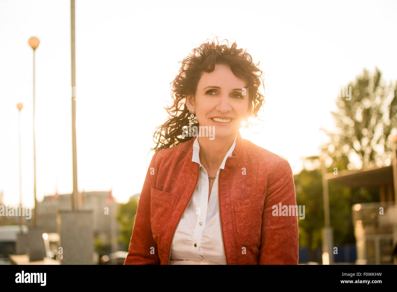 Ritratto di sorridente donna senior indossando giacca rossa in strada con Sun in background Foto Stock