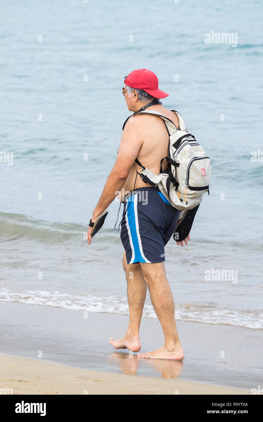 Uomo anziano camminando sulla spiaggia in Spagna con il berretto da baseball sulla parte posteriore a quella anteriore Foto Stock