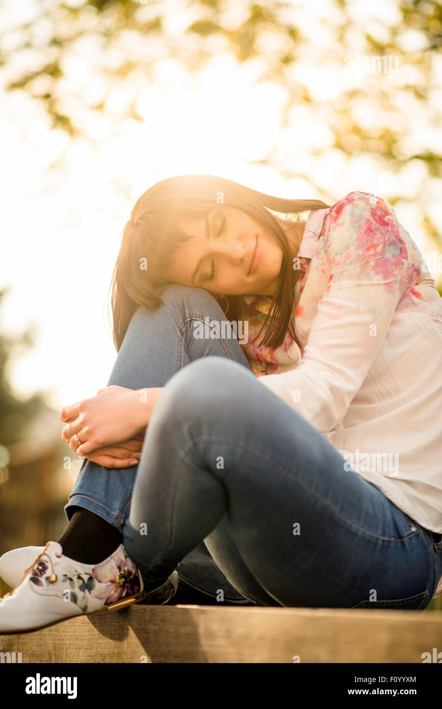 Ritratto di donna appoggiata sul suo ginocchio con gli occhi chiusi seduta sul banco al tramonto Foto Stock