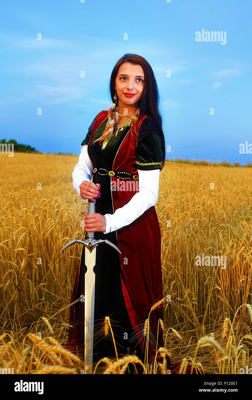Sorridente giovane donna con abito ornamentali e spada in mano in piedi su un campo di grano con il tramonto del sole. Sfondo naturale.. Foto Stock