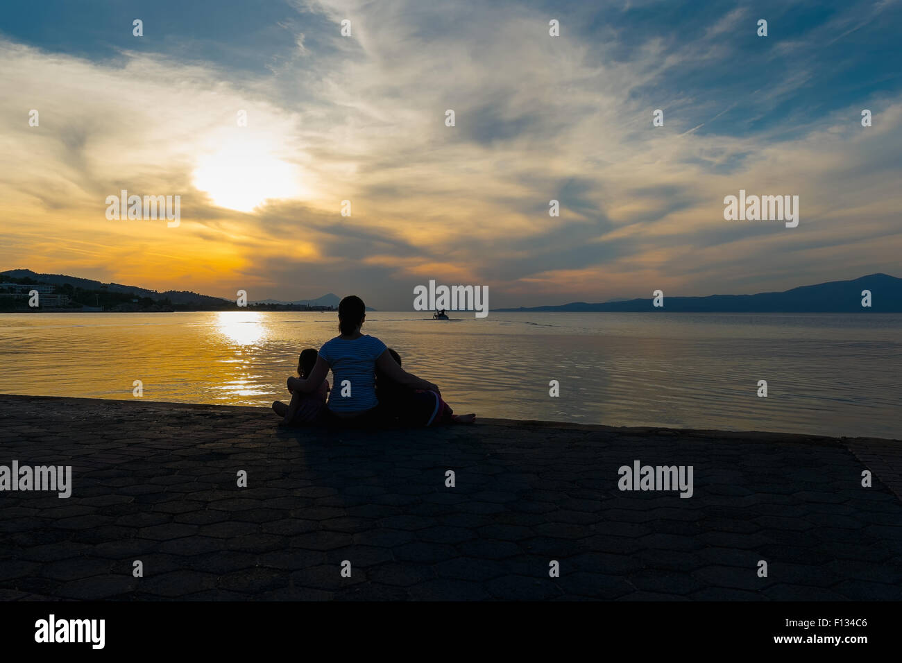 Madre e le sue figlie e guardare il tramonto e la silhouette di una barca da pesca.Una famiglia amorevole momento contro un cielo drammatico. Foto Stock