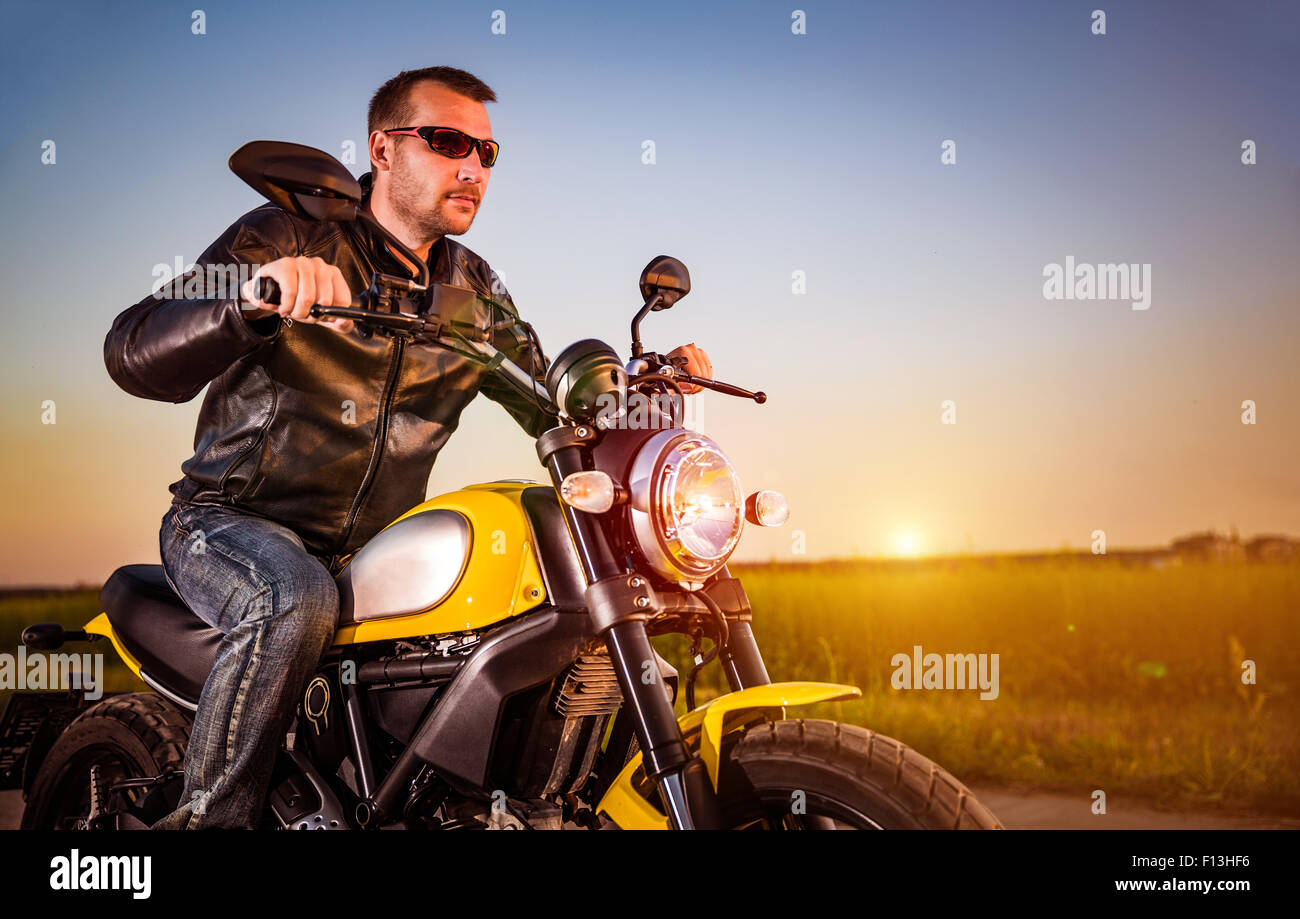 Biker uomo che indossa una giacca di pelle e occhiali da sole seduto sulla sua moto guardando il tramonto. Foto Stock Biker uomo che indossa una giacca di pelle e occhiali da sole seduto sulla sua moto guardando il tramonto. Foto Stock