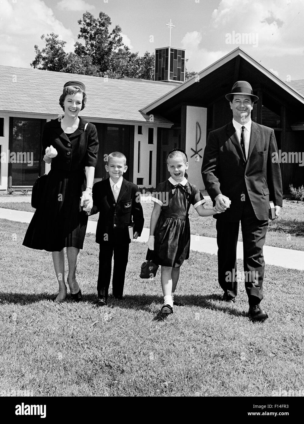 Anni sessanta sorridente mano azienda famiglia di quattro padre madre figlio FIGLIA A PIEDI DALLA CHIESA guardando la fotocamera Foto Stock
