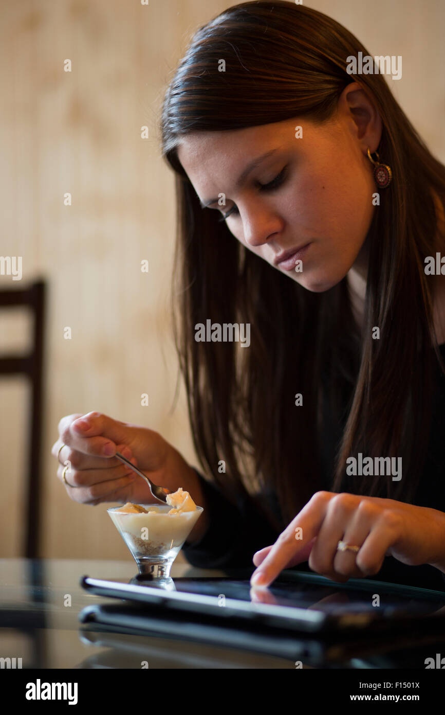Grave donna con capelli lunghi marrone a mangiare il gelato e con tavoletta digitale a tavola Foto Stock
