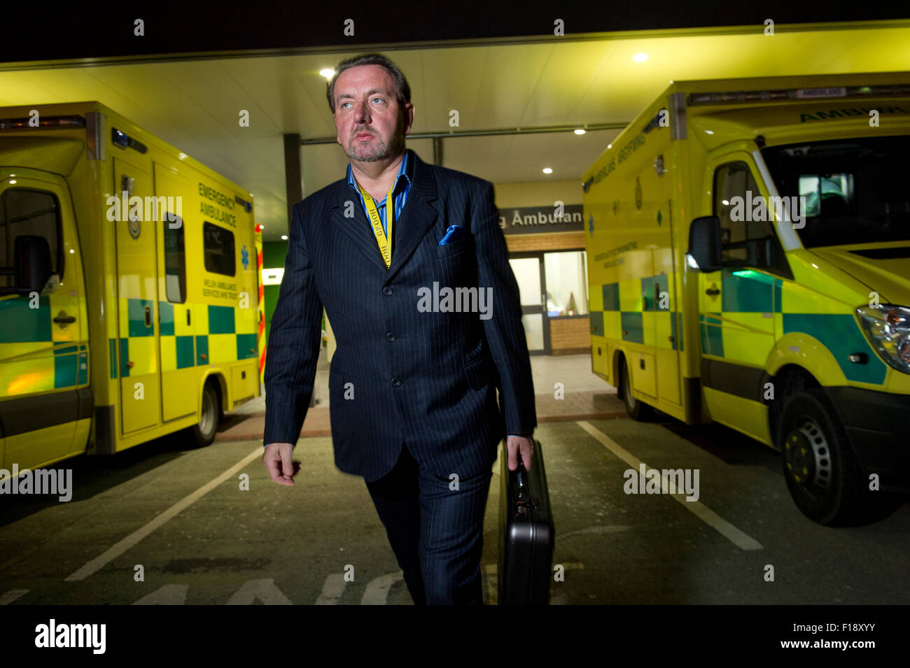 Un maschio fuori orario, dottore su chiamata, tenendo medical bag e del telefono mobile, a camminare lungo una linea di ambulanze presso un ospedale UK GP Foto Stock