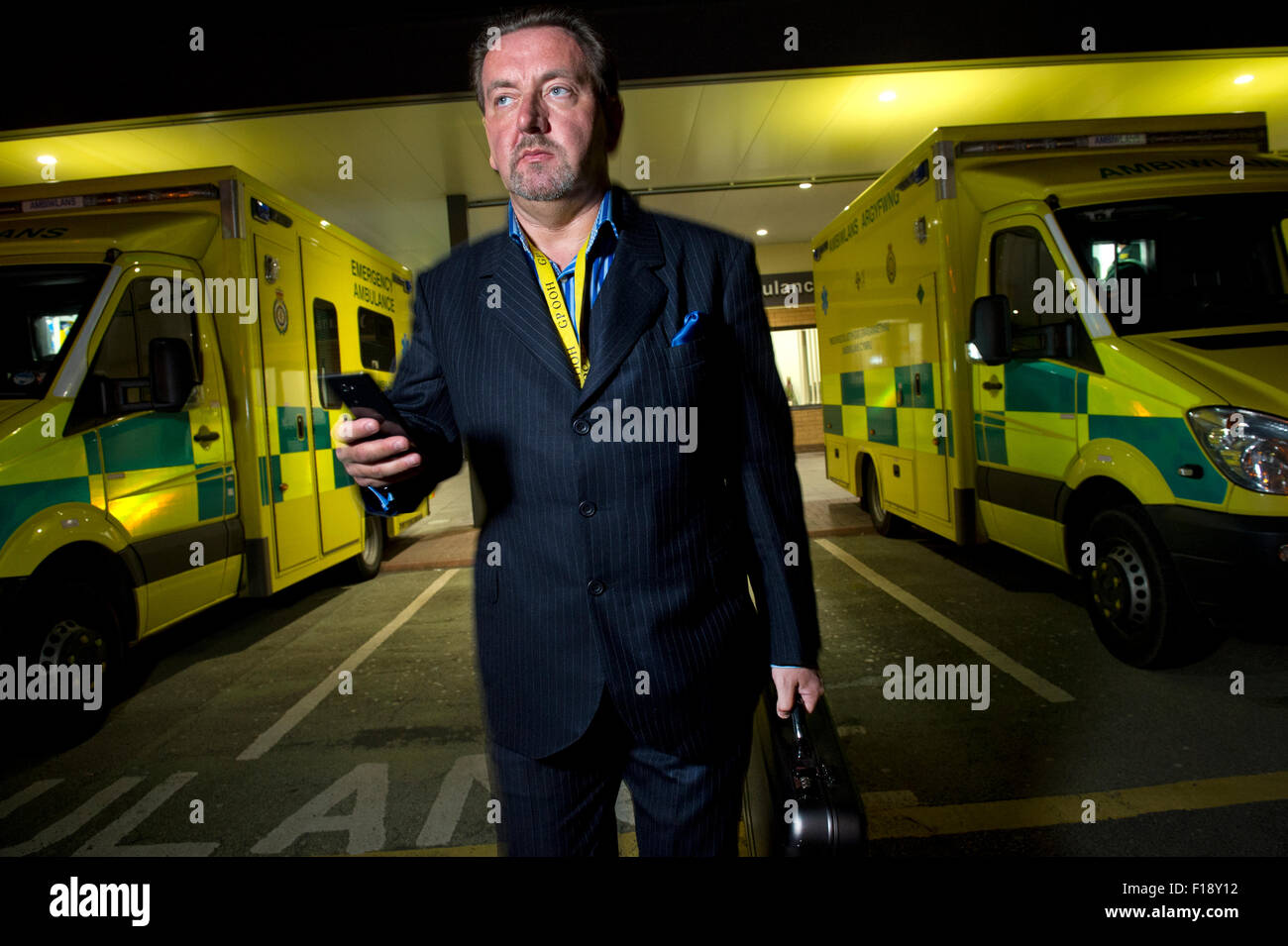 Un maschio fuori orario, dottore su chiamata, tenendo medical bag e del telefono mobile, a camminare lungo una linea di ambulanze presso un ospedale UK GP Foto Stock