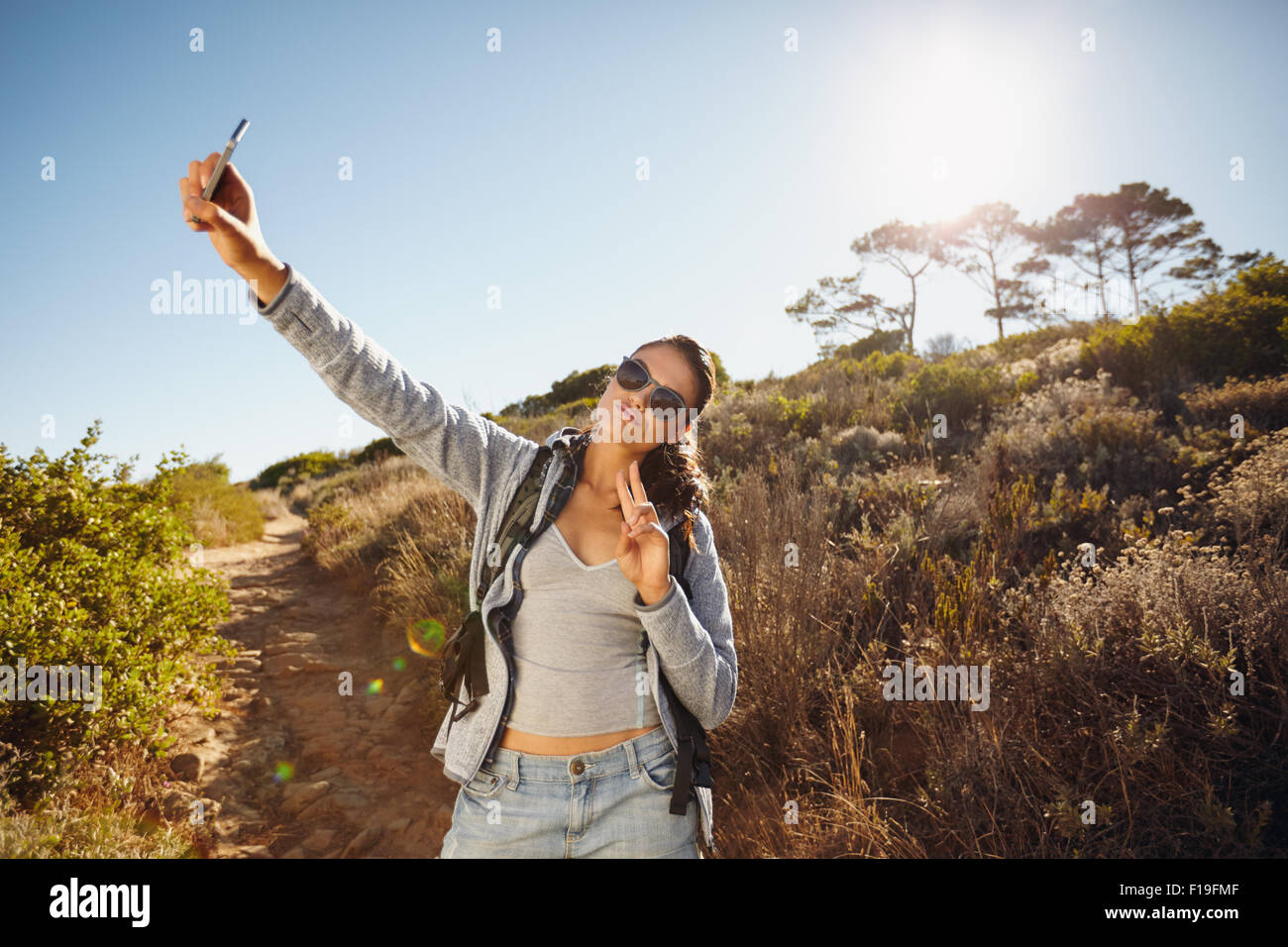 Felice ed energico giovane donna prendendo un selfie. Lei tiene la fotocamera del cellulare ad alta in posa e gesticolando vittoria il segno w Foto Stock