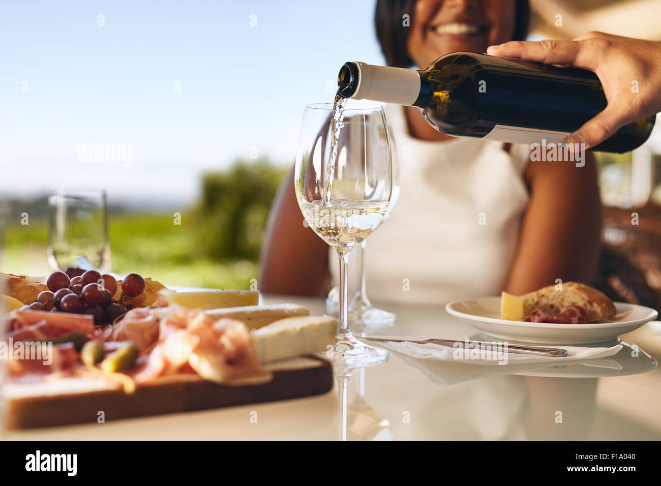 Le mani di un uomo versando il vino bianco in due bicchieri da una bottiglia con una donna sorridente in background in cantina. Focus su vetri e Foto Stock