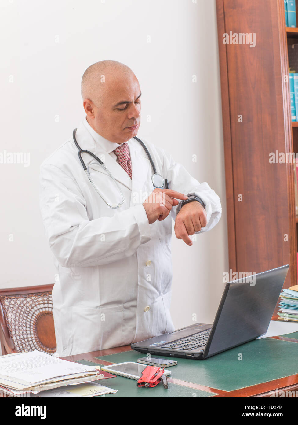 Sconcertato medico nel suo studio, utilizza un smartwacth davanti al suo computer portatile. Utilizzare le nuove tecnologie. Nel suo studio professionale, egli è sitti Foto Stock