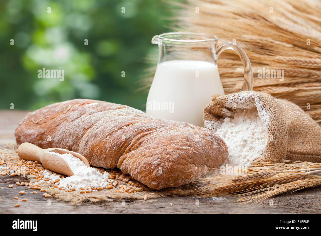 Pane fresco, brocca del latte, sacco di farina di grano e un covone di grano su un tavolo di legno. Foto Stock