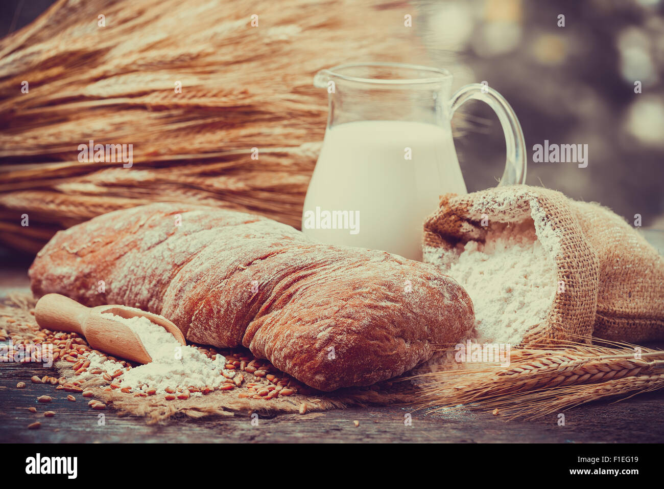 Pane fresco, brocca del latte, sacco di farina di grano e un covone di grano su un tavolo di legno. Retrò stilizzato. Foto Stock