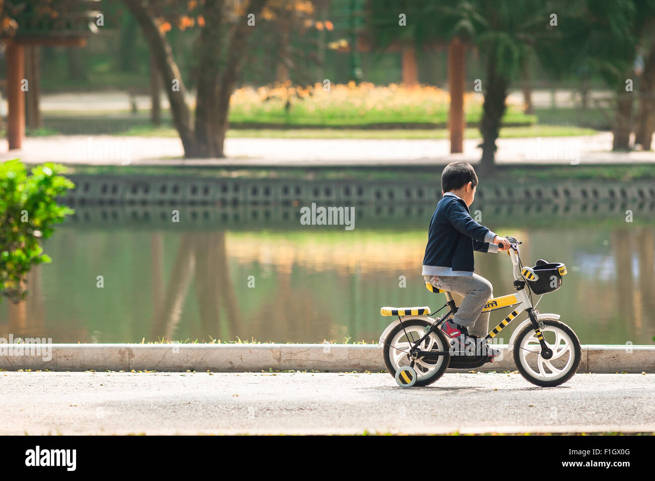 Bambini che giocano da soli, veduta di un bambino in bicicletta davanti a un lago nel Giardino Botanico di Hanoi, Vietnam. Foto Stock