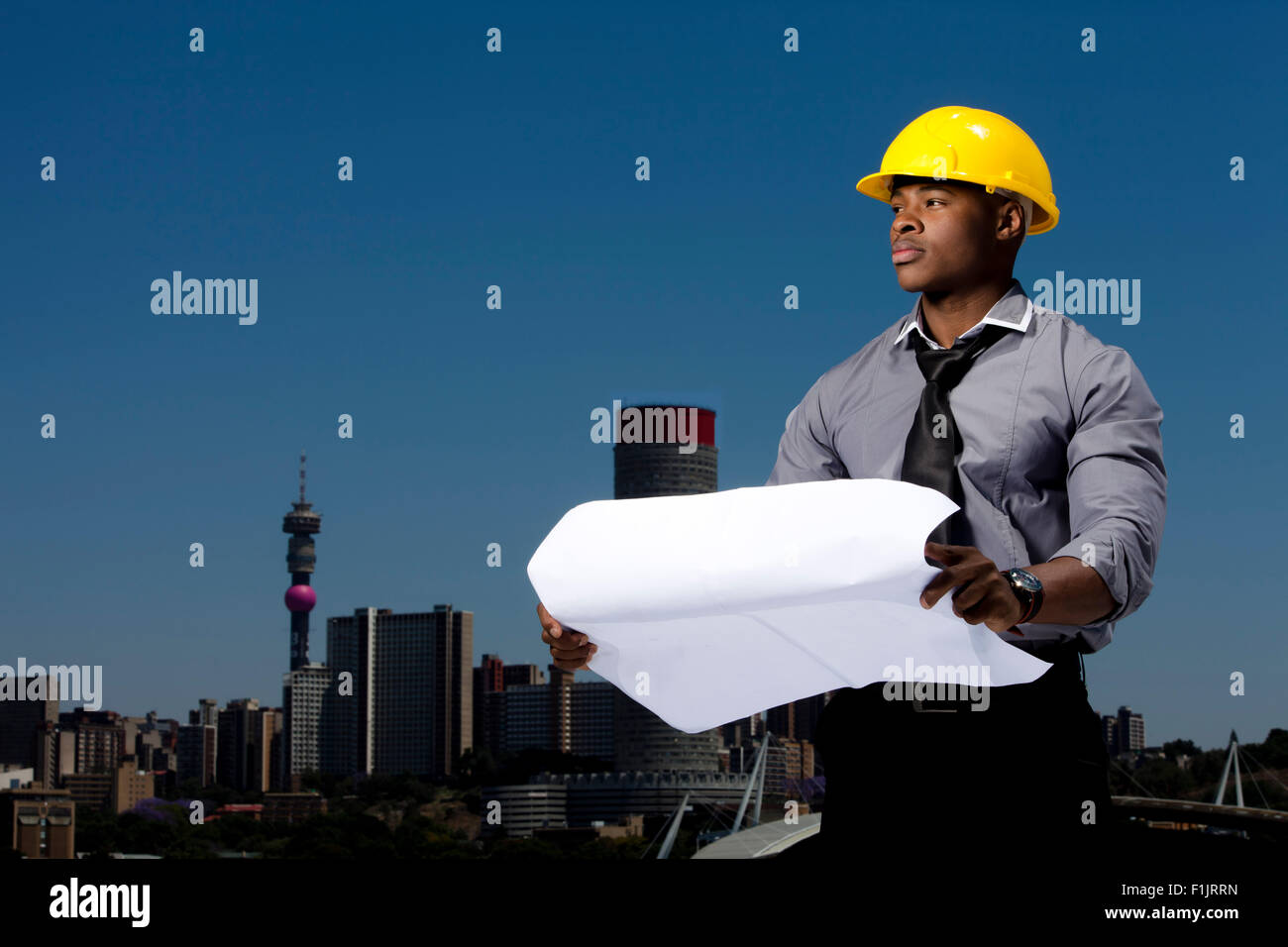 Elegantemente vestito uomo d affari indossando hardhat detiene i progetti di un edificio mentre in piedi con le braccia incrociate con cityscape in background Foto Stock