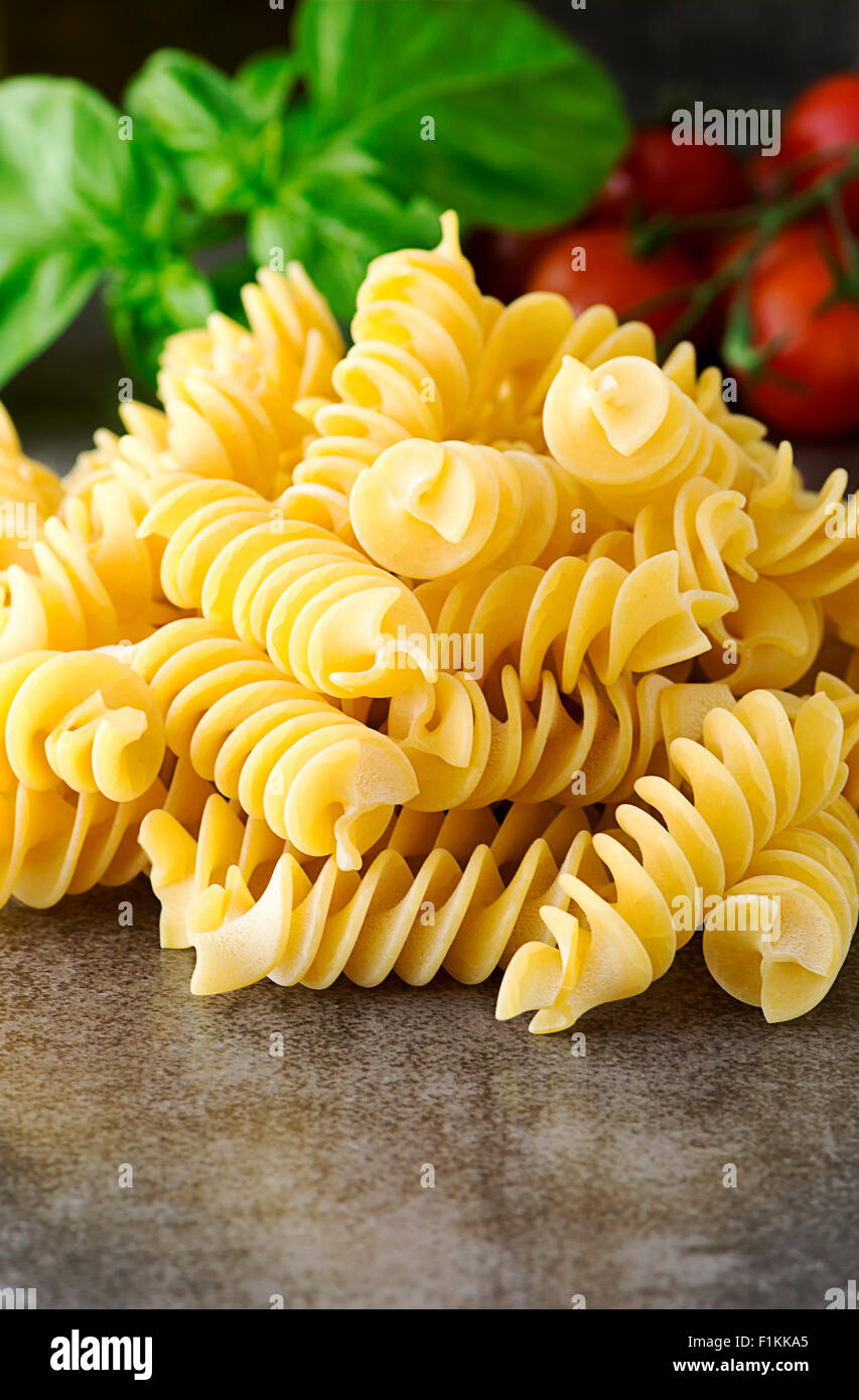Fusilli non cotti fotografato con il fuoco selettivo nella finestra luce su un effetto di ardesia piastrella con basilico e pomodorini Foto Stock