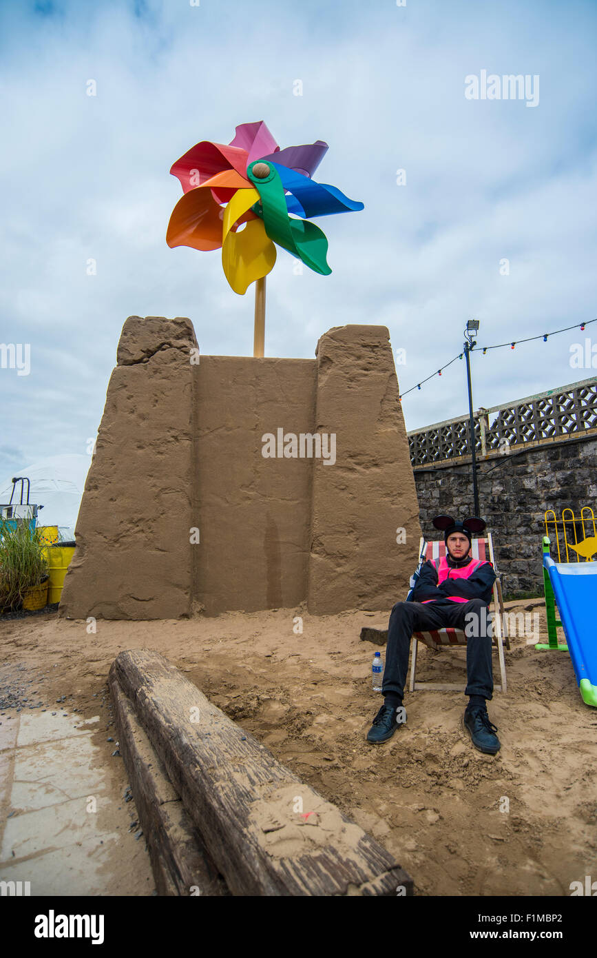 Un dipendente di Banksy's Dismaland Bemusement Park si siede su una finta spiaggia di fronte a un enorme sandcastle. Foto Stock