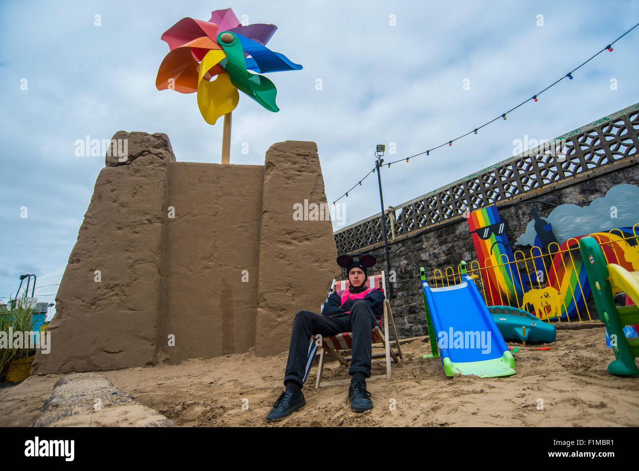 Un dipendente di Banksy's Dismaland Bemusement Park si siede su una finta spiaggia di fronte a un enorme sandcastle. Foto Stock