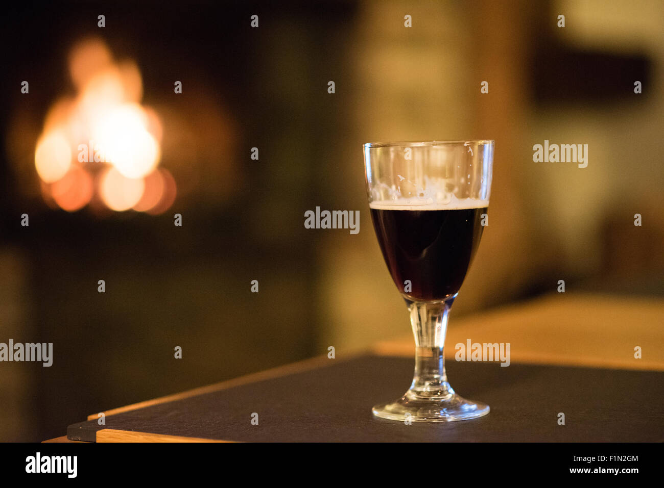 Piccolo singolo bicchiere di birra scura su un tavolo di fronte un accogliente il fuoco reale su un inverno di pomeriggio in Inghilterra Foto Stock