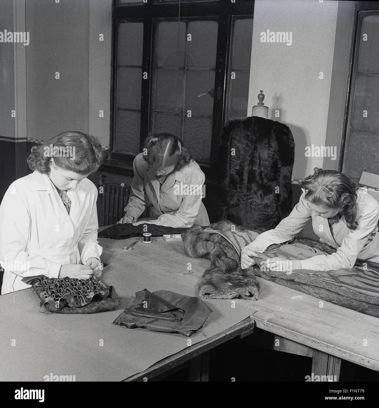 anni '1950, tre donne sartoriali o apprendiste su un banco da lavoro per rifinire o riparare un cappotto di pelliccia da donna. Un altro cappotto di pelliccia su un piedistallo nella stanza. Foto Stock