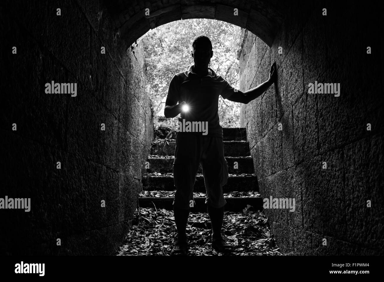 Giovane con una torcia elettrica entra nel tunnel di pietra e guarda al buio, foto in bianco e nero Foto Stock