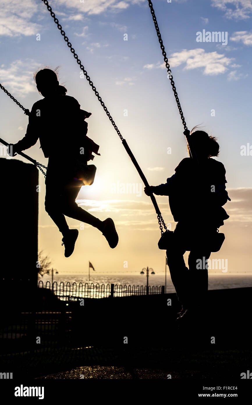 Silhouette di due donne bambini che giocano sulle altalene contro un cielo blu con il sole inizio per impostare Foto Stock
