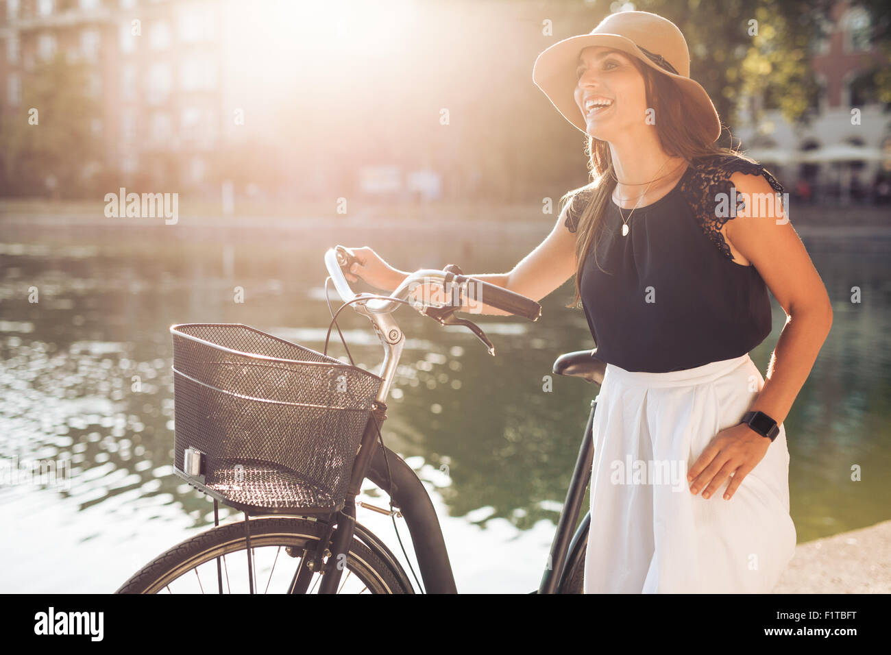 Ritratto di giovane e bella donna che indossa un cappello con una bicicletta a piedi lungo un laghetto. Donna felice con una bicicletta presso il parco cercando Foto Stock