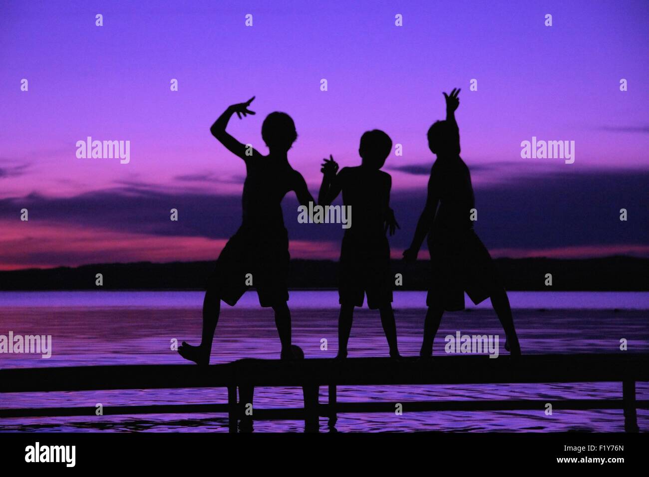 Tre sagome di ragazzi su un dock contro un tramonto Foto Stock