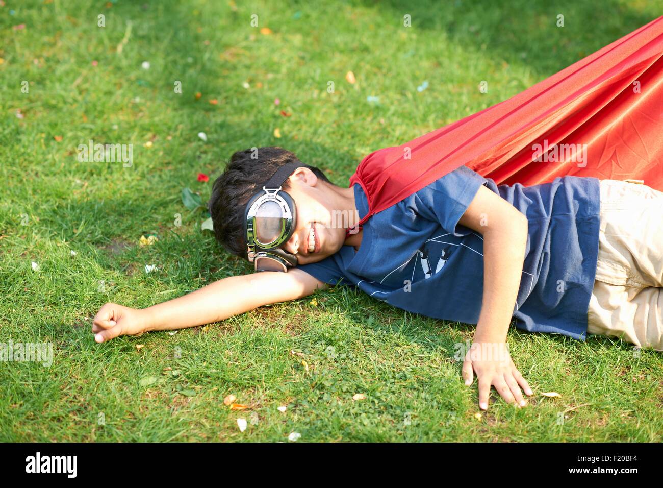Ragazzo che indossa gli occhiali e cape giacente sul lato su erba, atteggiamento di volo Foto Stock