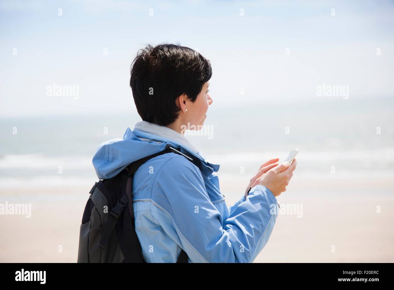 Metà donna adulta scrivere messaggi su smartphone da spiaggia mentre guardando al mare Foto Stock