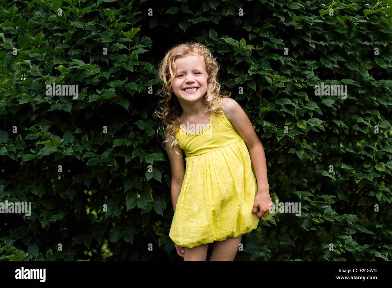 Ritratto di bionda sorridente ragazza dai capelli indossando abito giallo in piedi nella parte anteriore del giardino hedge Foto Stock