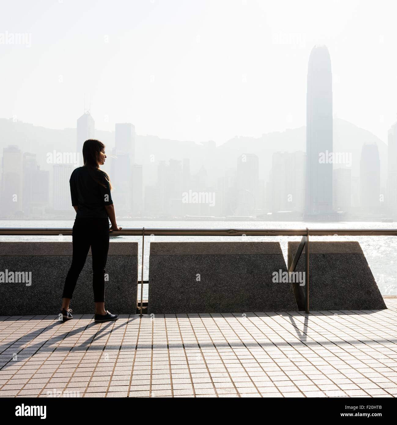 Stagliano vista laterale della giovane donna in piedi che guarda fuori acqua oltre a skyline di Hong Kong, Cina Foto Stock