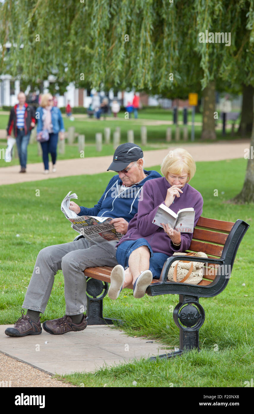 Coppia di mezza età rilassante, la signora con un libro in brossura e l'uomo con un quotidiano in una sede del parco, England, Regno Unito Foto Stock