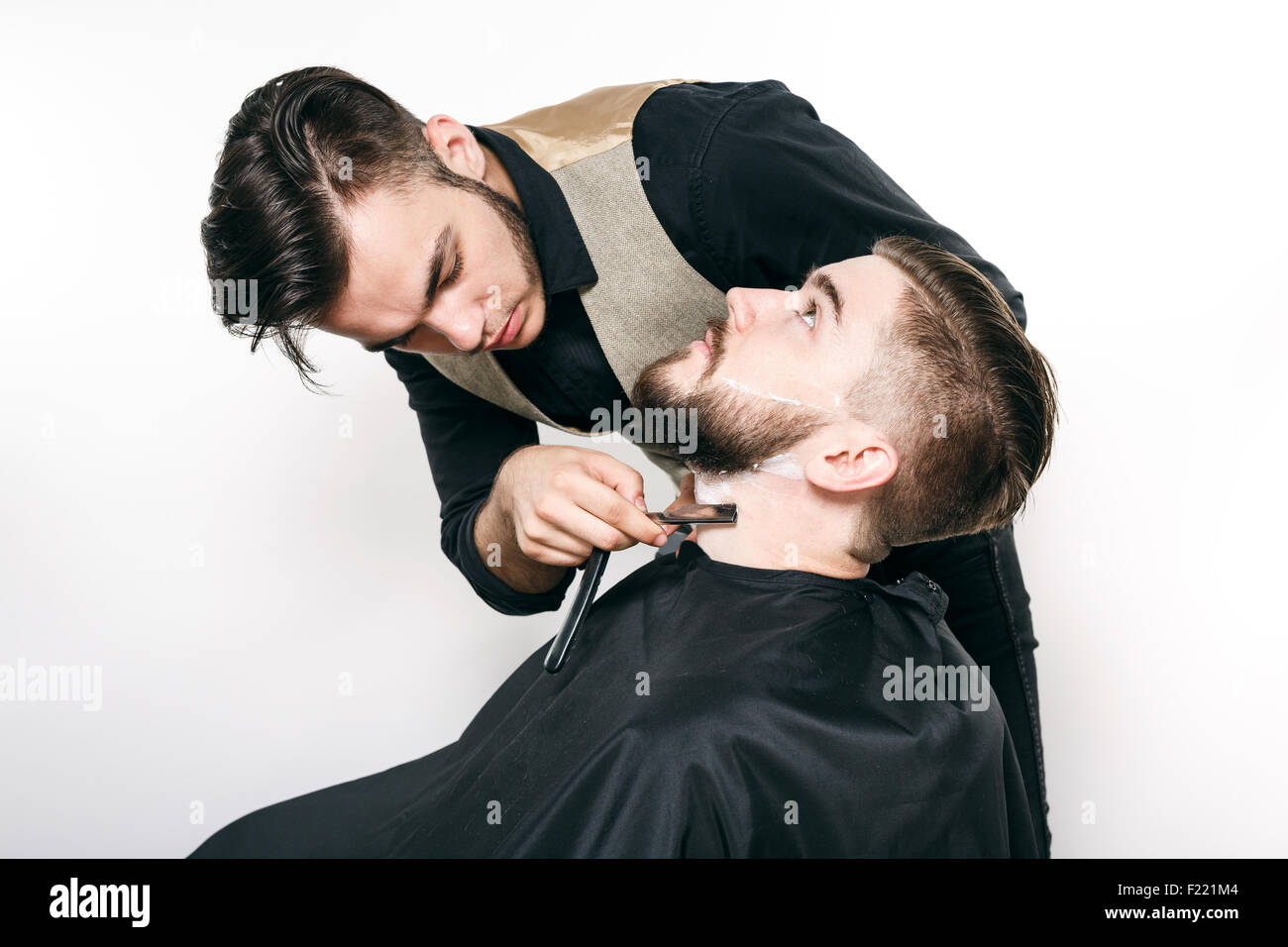 Brunette barbiere, indossa in camicia nera e il panciotto, rasatura barbuto cliente con rasoio, su sfondo bianco, in studio, WAIS Foto Stock