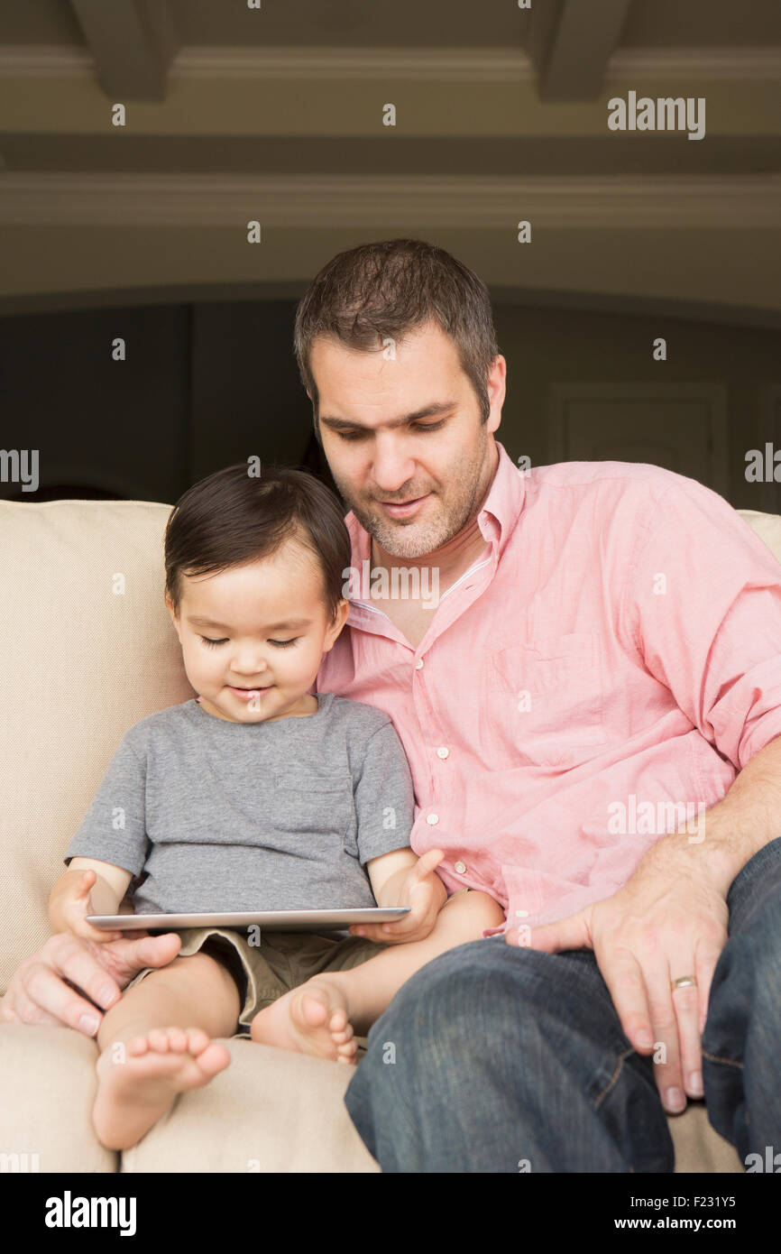 Uomo sorridente seduto su un divano con il suo giovane figlio, guardando una tavoletta digitale. Foto Stock