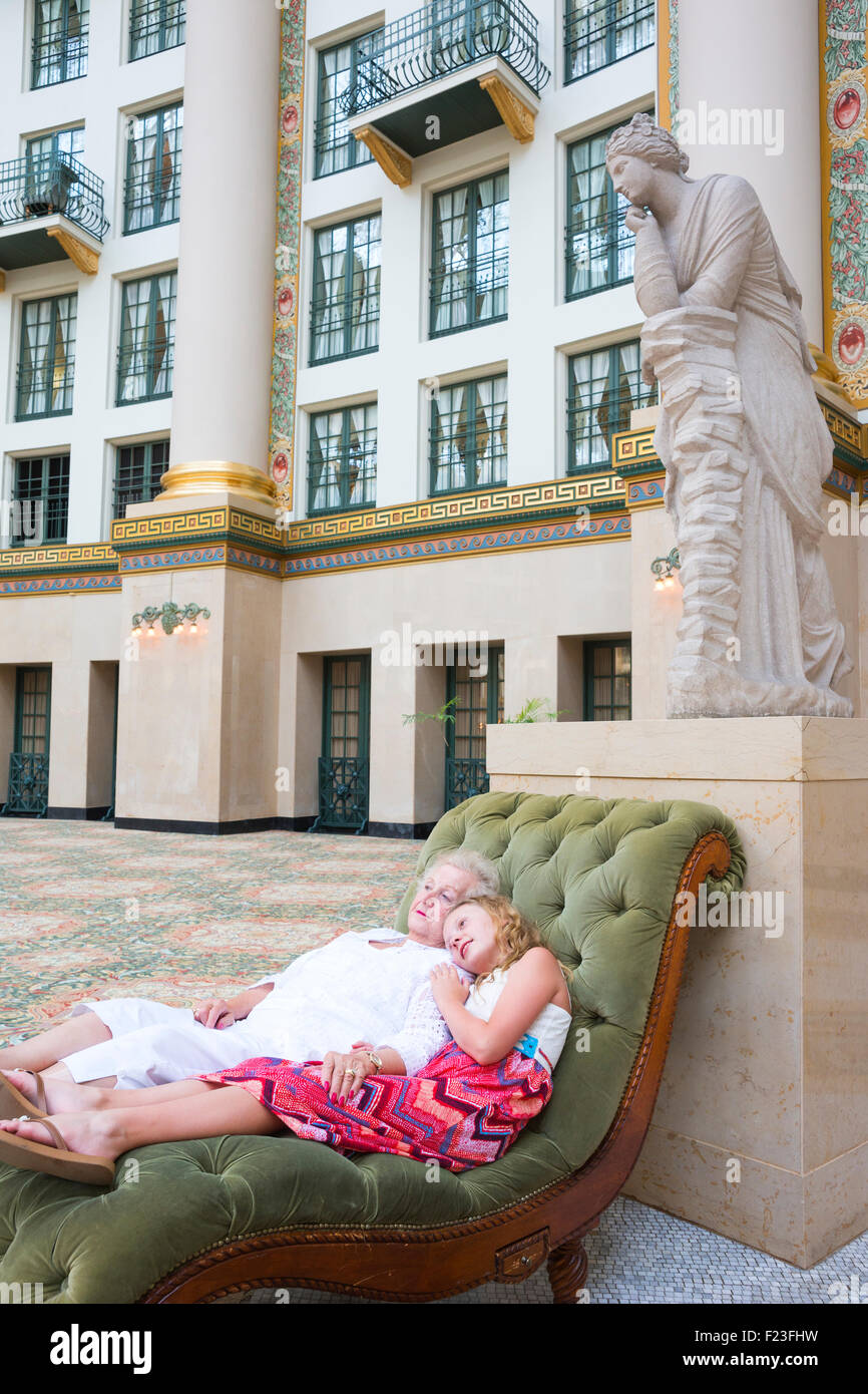 Senior caucasica donna e suo nipote che indossa un abito rosa sdraiati su una chaise longue velluto sotto una statua in marmo Foto Stock