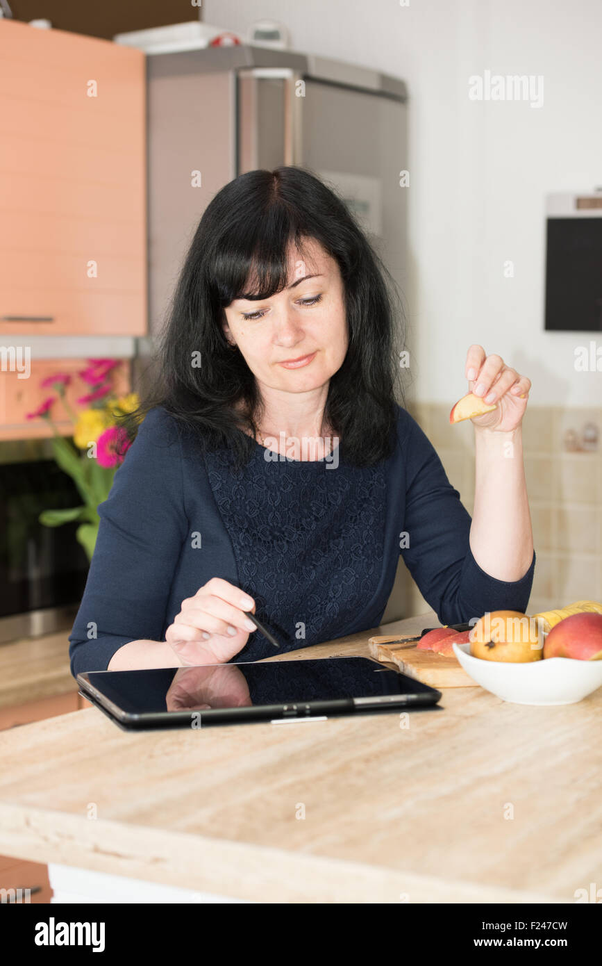 Donna felice mangiare frutta e utilizzo di tablet pc in cucina Foto Stock