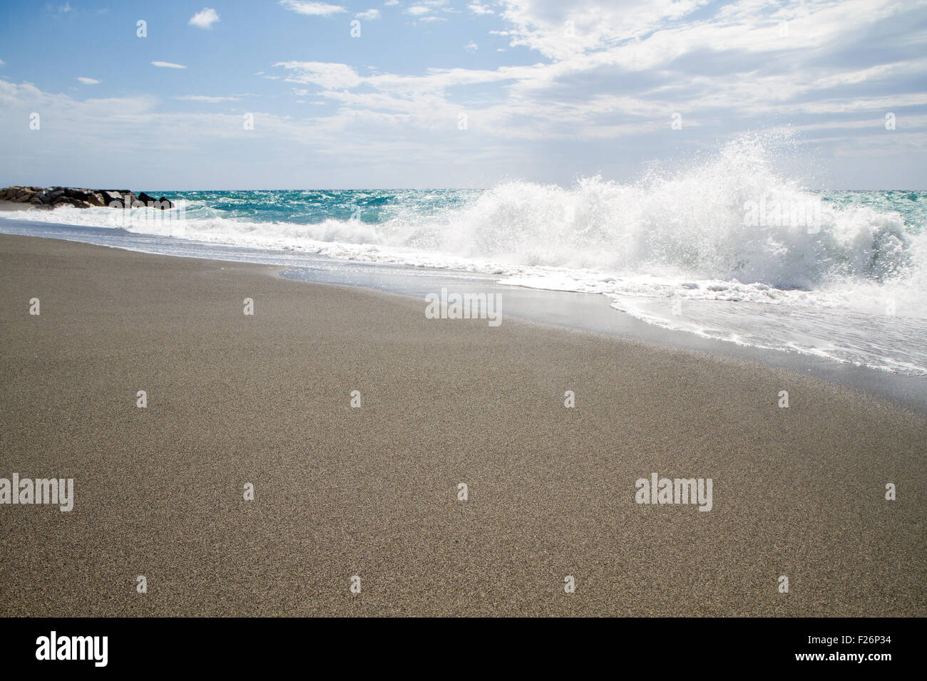 Le onde che si infrangono sulla spiaggia deserta, sullo sfondo azzurro del cielo e delle nubi Foto Stock