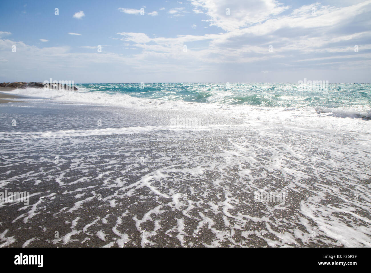 Le onde che si infrangono sulla spiaggia deserta, sullo sfondo azzurro del cielo e delle nubi Foto Stock