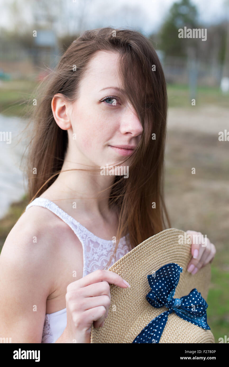 Freckled felice ragazza in abito bianco azienda cappello di paglia nelle sue mani e guardando la fotocamera in tempo ventoso Foto Stock