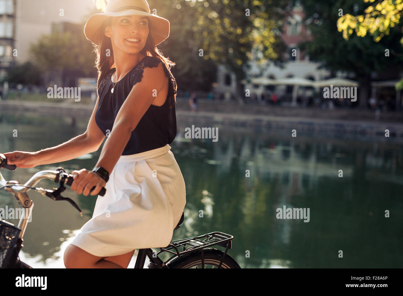 Ritratto di felice giovane femmina la bicicletta da un laghetto che guarda lontano sorridente. Donna che indossa un cappello su un giorno di estate in sella la sua bicicletta. Foto Stock