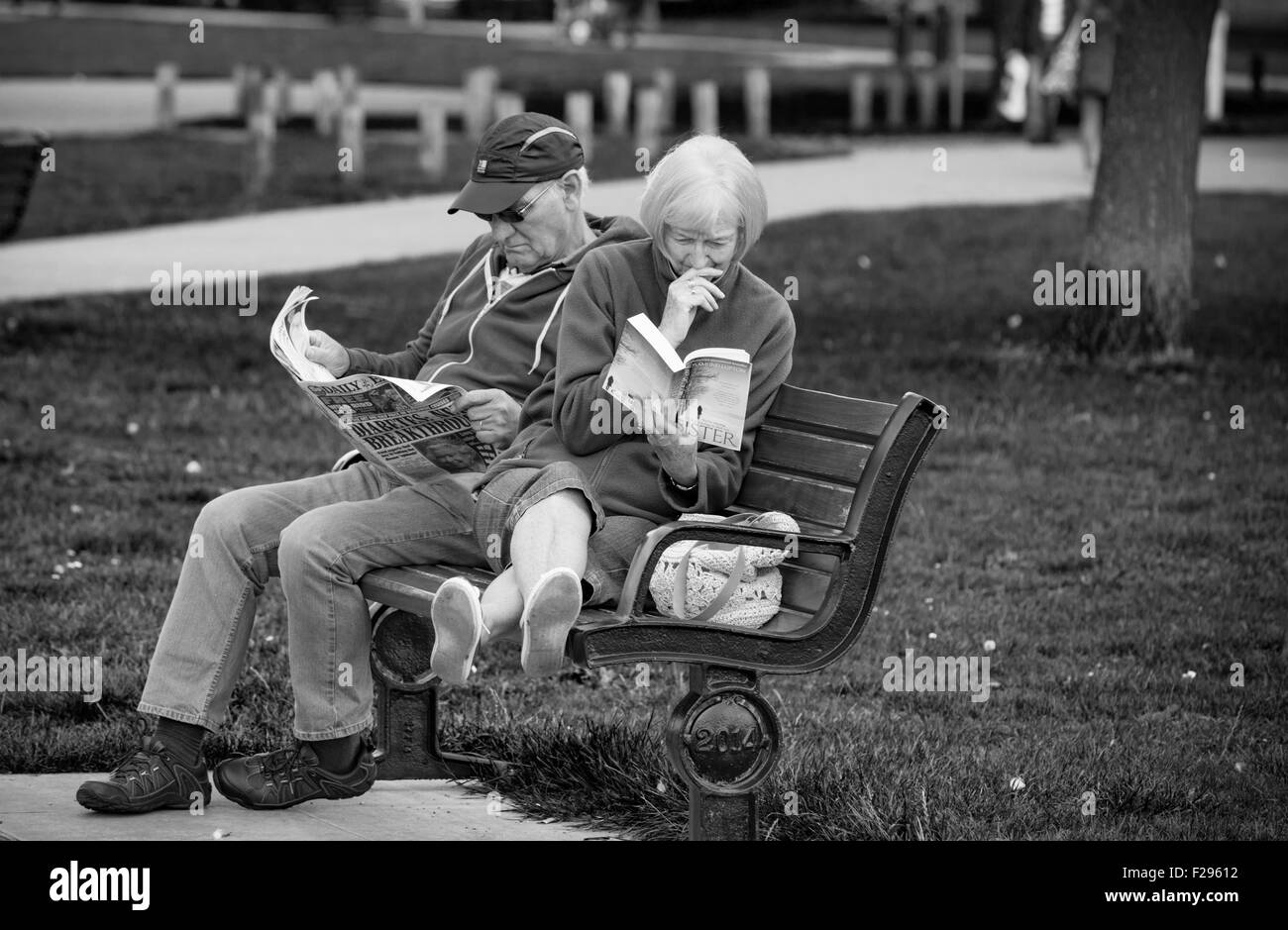 Coppia di mezza età rilassante, la signora con un libro in brossura e l'uomo con un quotidiano in una sede del parco, England, Regno Unito Foto Stock