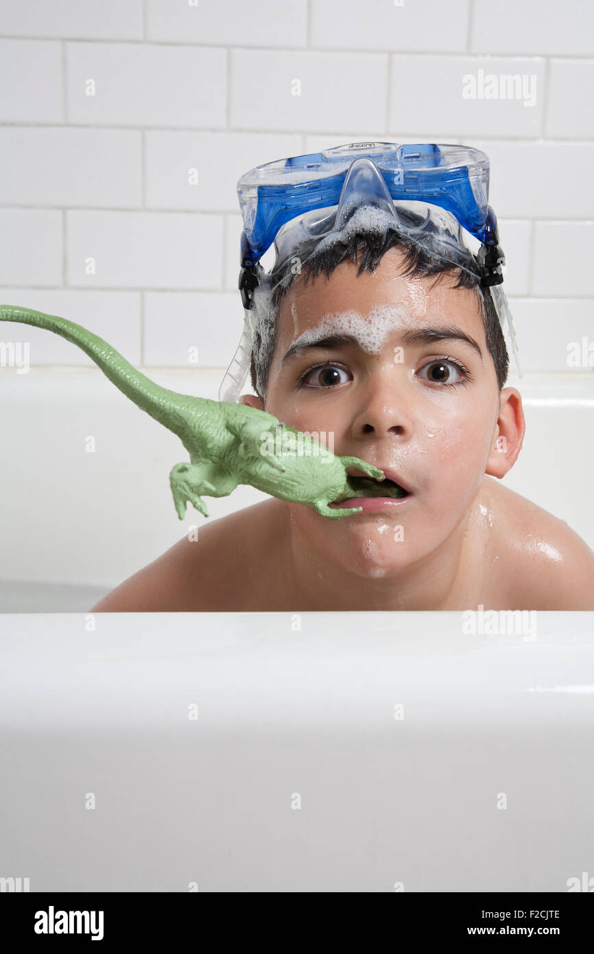 Giovane ragazzo in vasca con gli occhiali di protezione sulla cima della sua testa e dinosauro di plastica in bocca con schiuma di sapone guarda la fotocamera Foto Stock