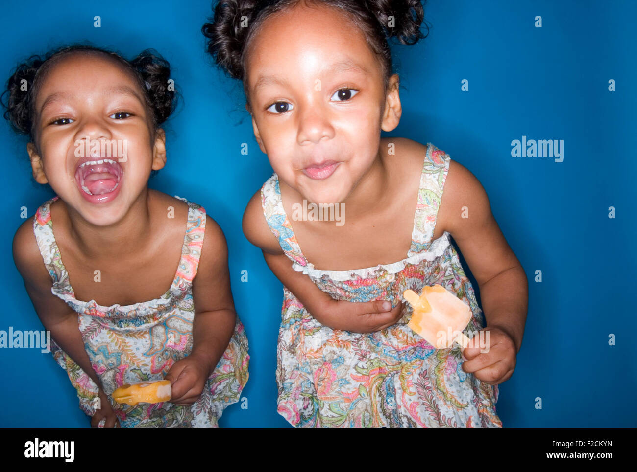 Due giovani gemello ragazze mangiare popscicles arancione di fronte a uno sfondo blu Foto Stock