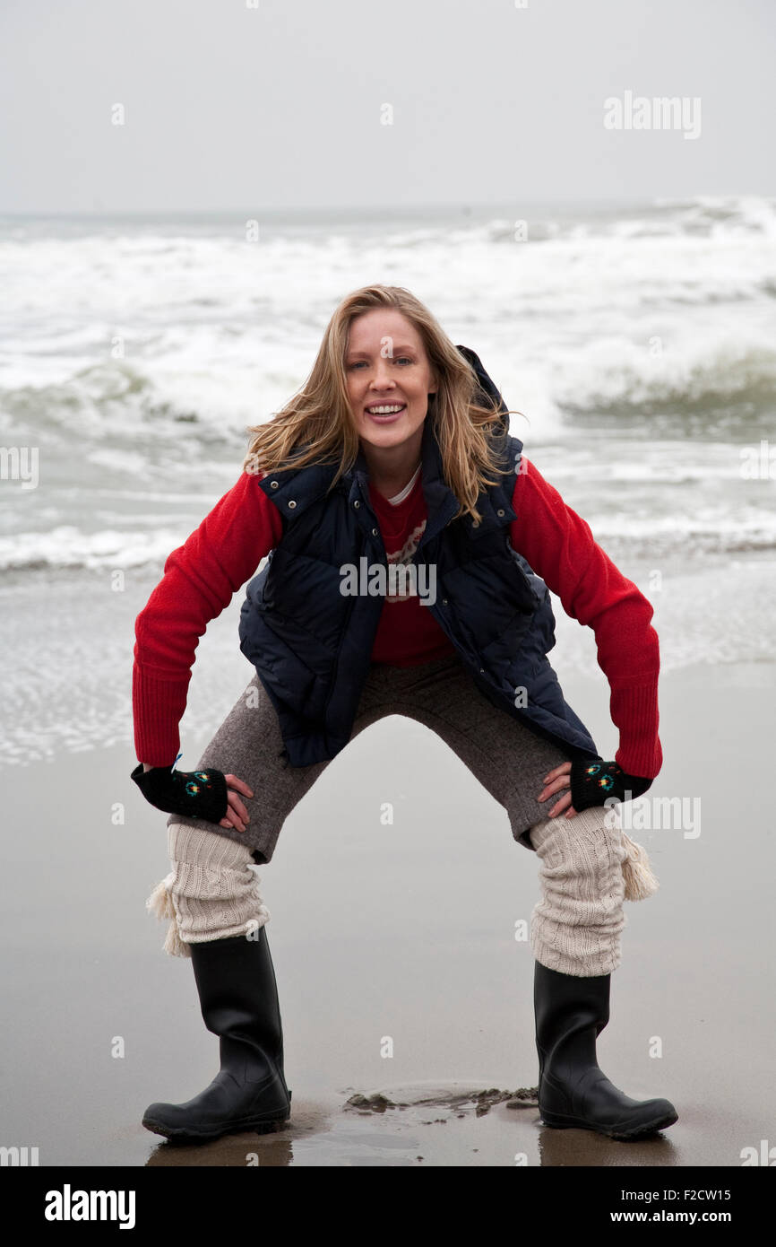 Giovane donna bionda si affaccia fotocamera, squatting in inverno sulla spiaggia di fronte oceano Foto Stock