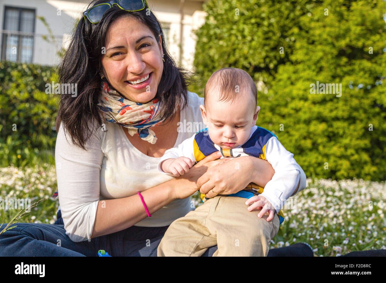 Carino 6 mesi baby con luce marrone capelli in bianco, blu e bruno camicia a maniche lunghe è abbracciato e tenuto da sua madre Foto Stock