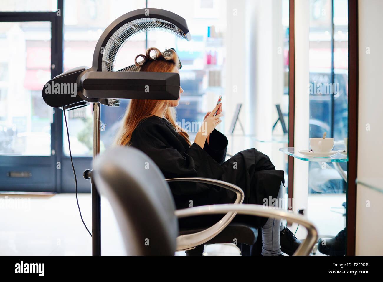 Donna seduta sotto essiccatore texting con un telefono cellulare nel salone di parrucchiere Foto Stock