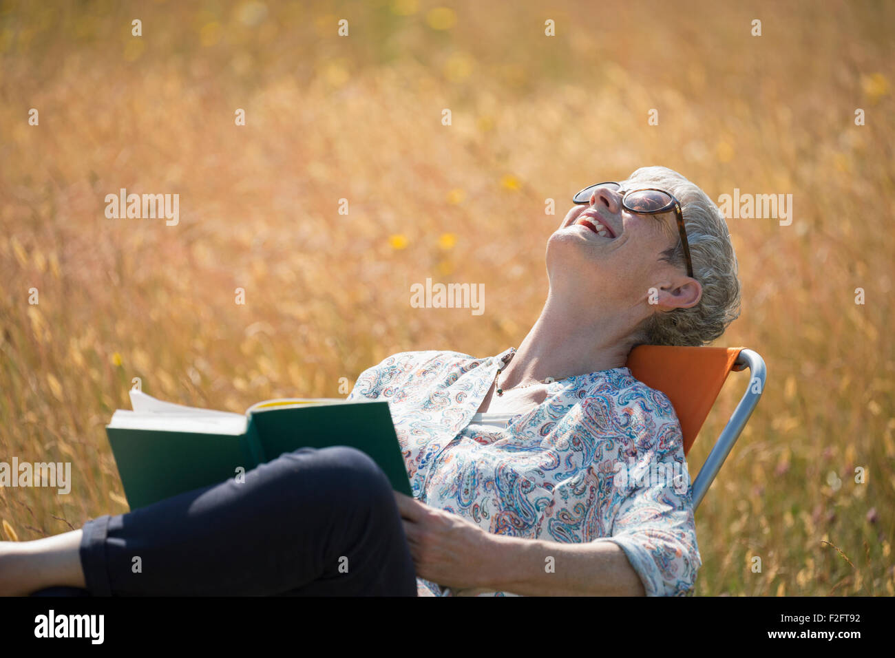 Senior donna libro di lettura e di ridere con la testa indietro nel campo soleggiato Foto Stock