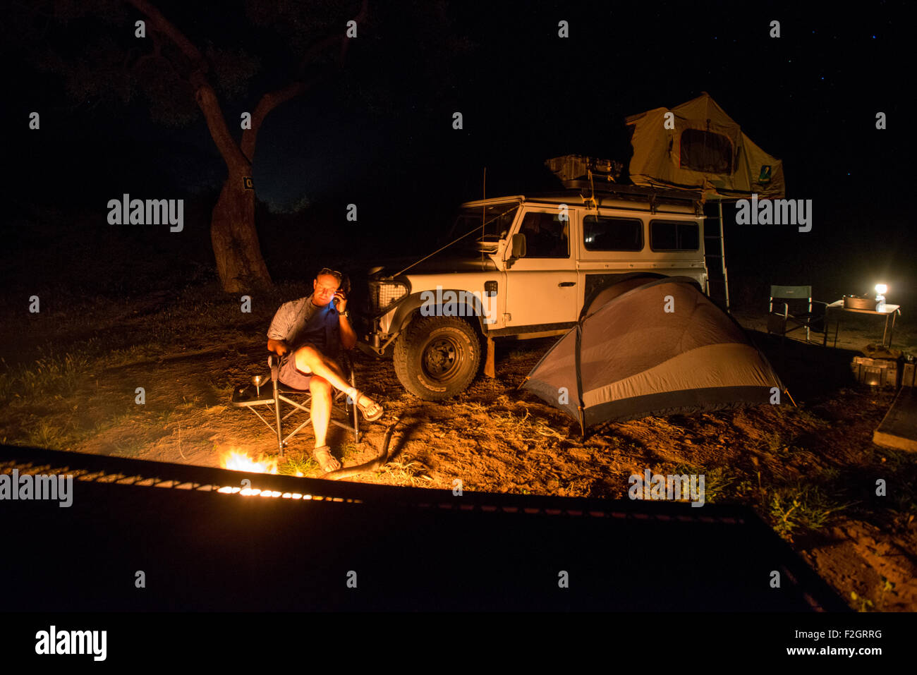 Uomo seduto in un incendio sul telefono con la sua Land Rover in background in Botswana, Africa Foto Stock