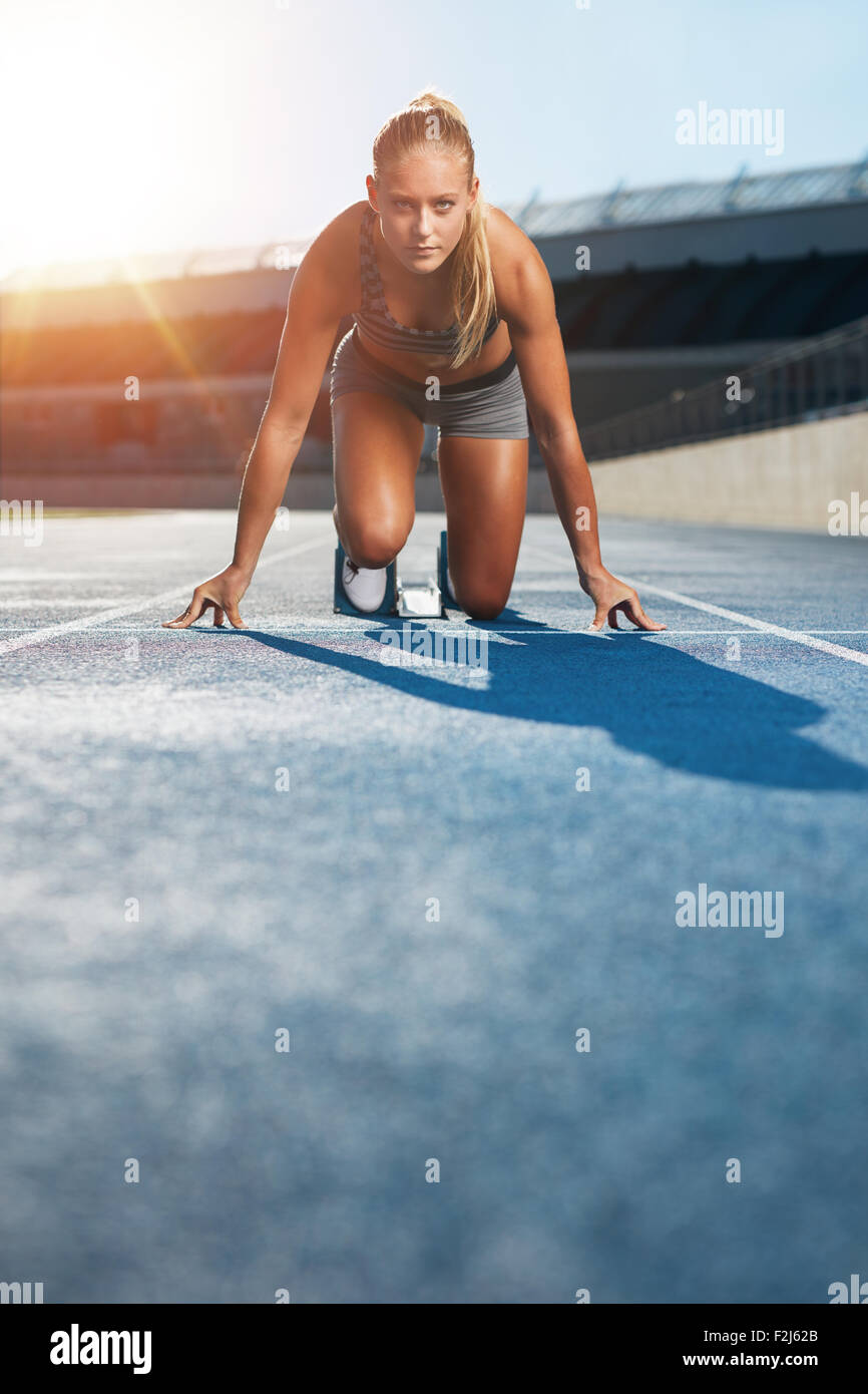 Giovane donna sprinter nella posizione avviamento su una pista a uno stadio sportivo cercando fino a telecamera con determinazione. Runner Foto Stock